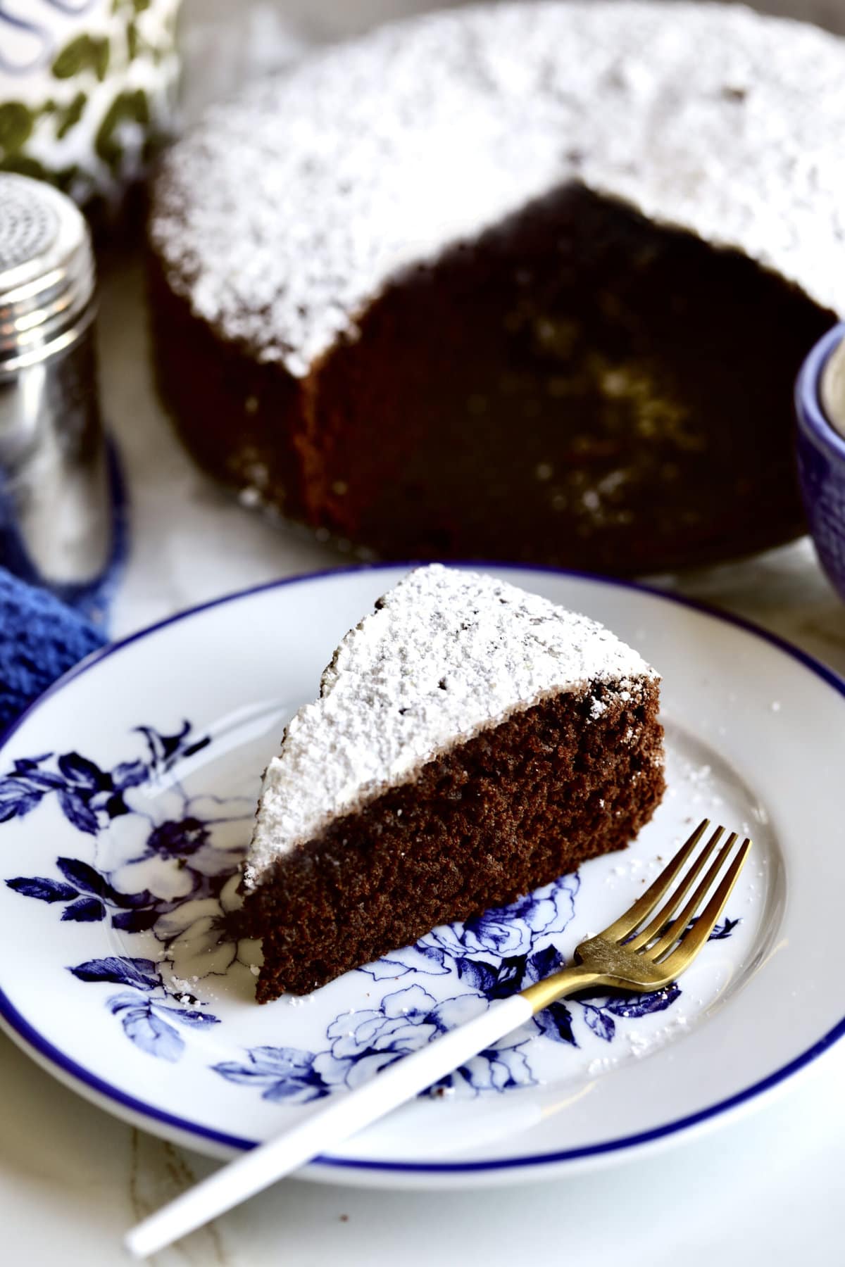 slice of chocolate olive oil cake on a plate with the whole cake in the background. 