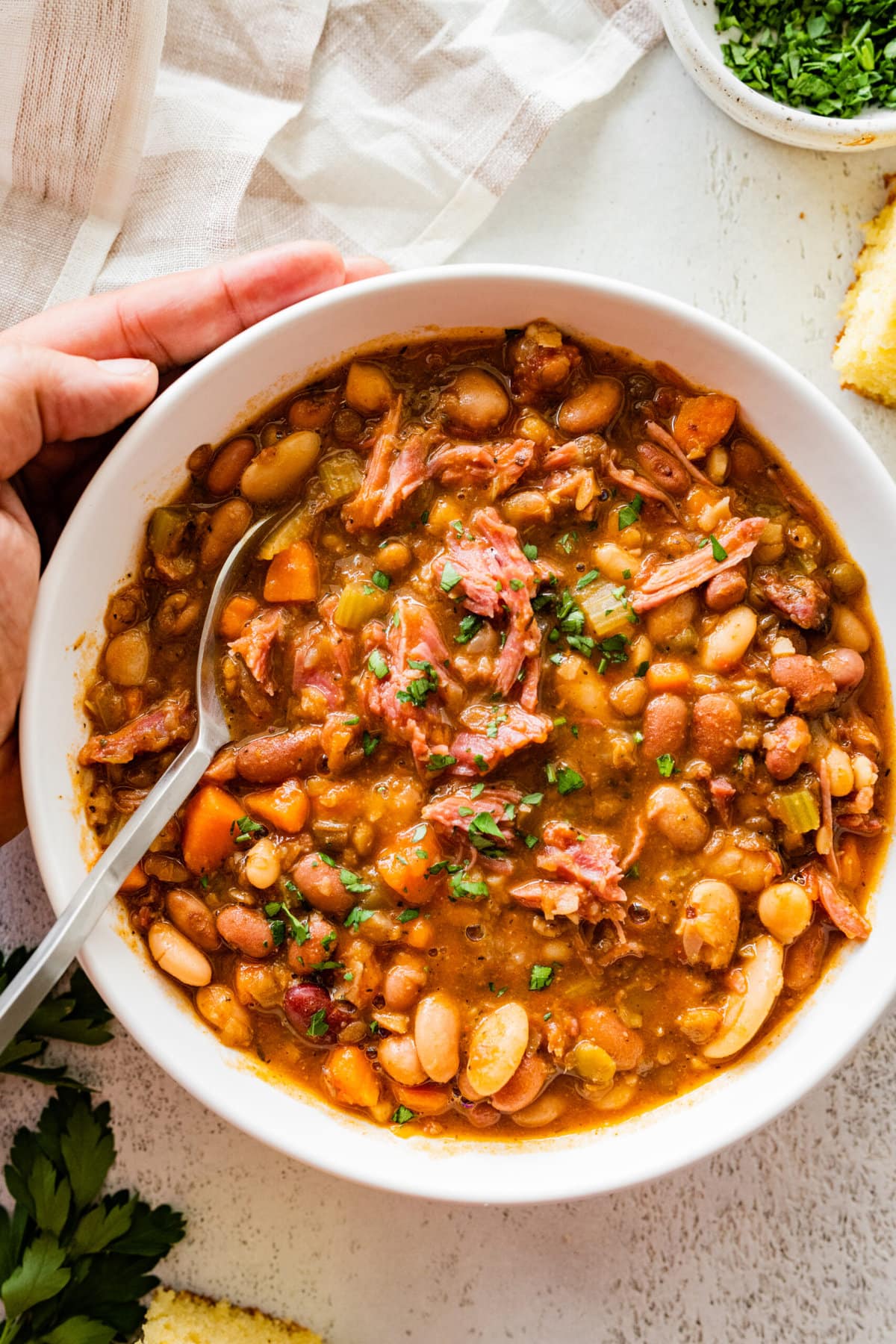 15 bean soup mix recipe in a white bowl with slices of cornbread on the side. Parsley for garnish. close up of bowl and soup.