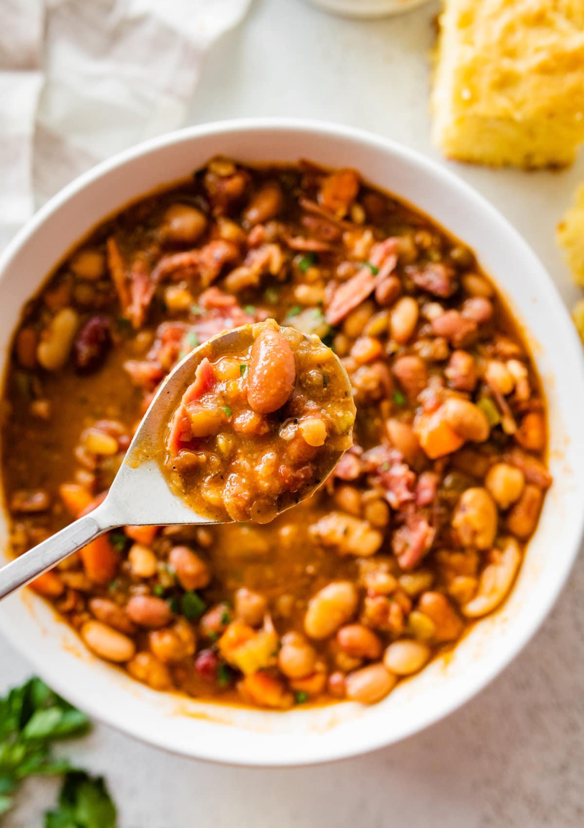 15 bean soup mix recipe in a white bowl with slices of cornbread on the side. Parsley for garnish. Spoon showing the soup.