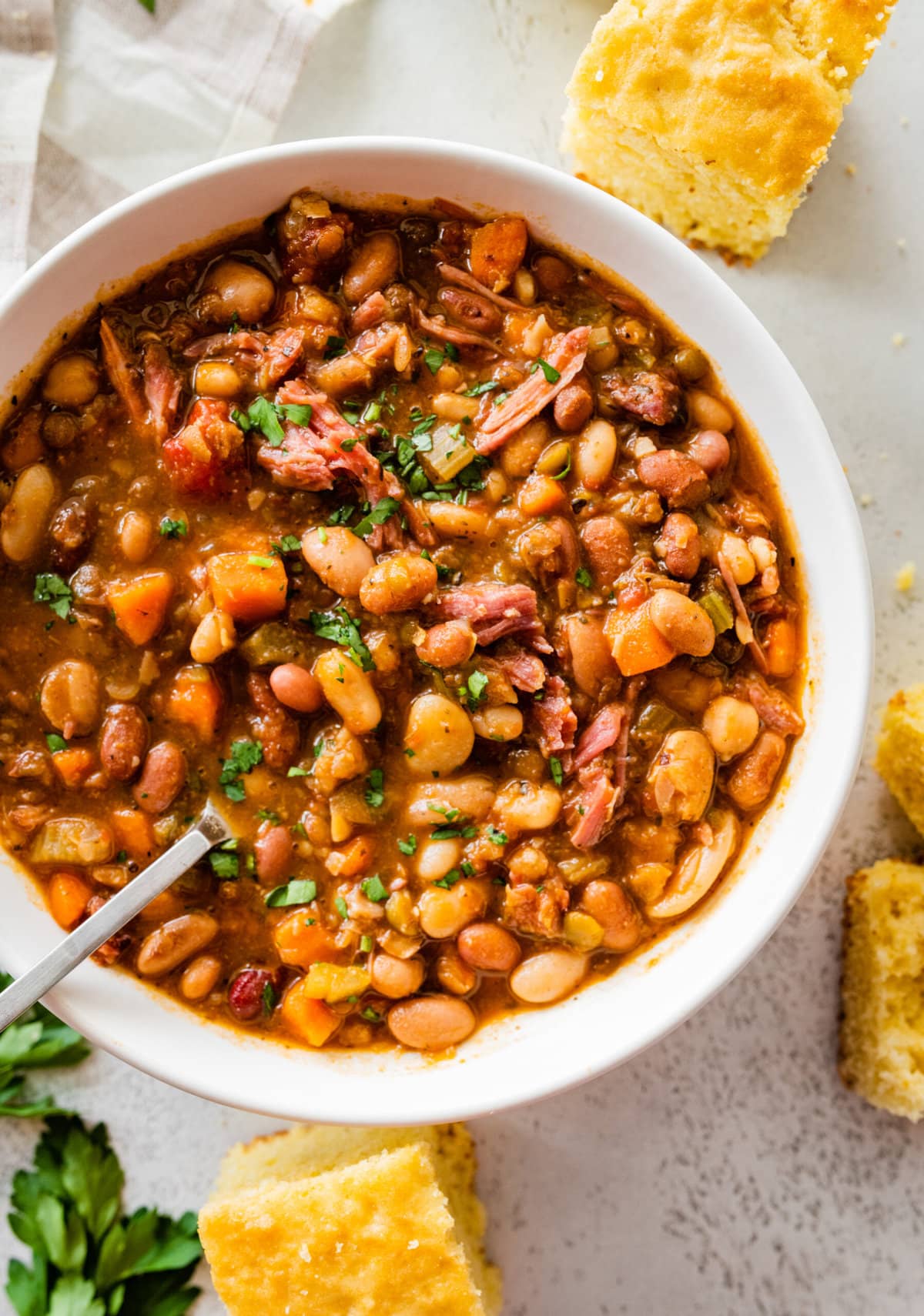 15 bean soup mix recipe in a white bowl with slices of cornbread on the side. Parsley for garnish.