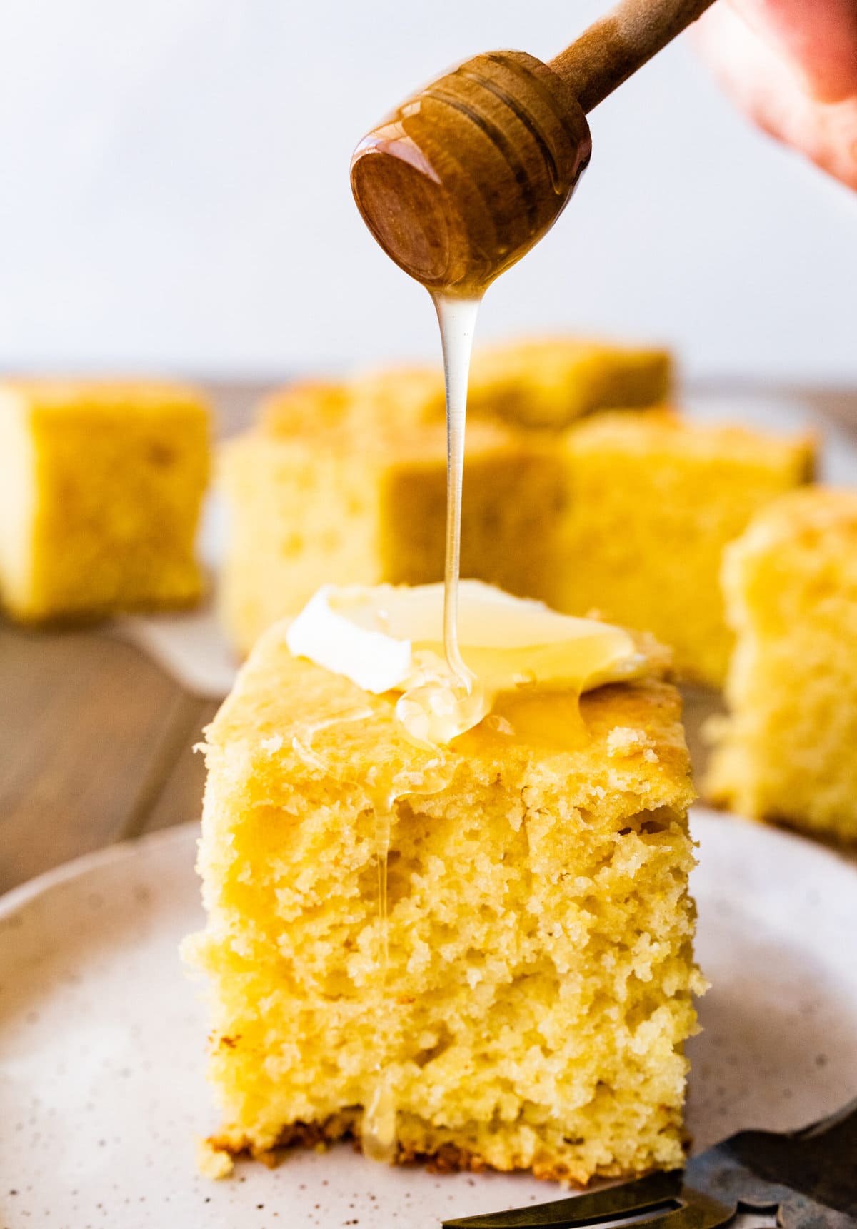 thick square of sweet cornbread on a plate with a pat of butter and honey drizzled on top. 