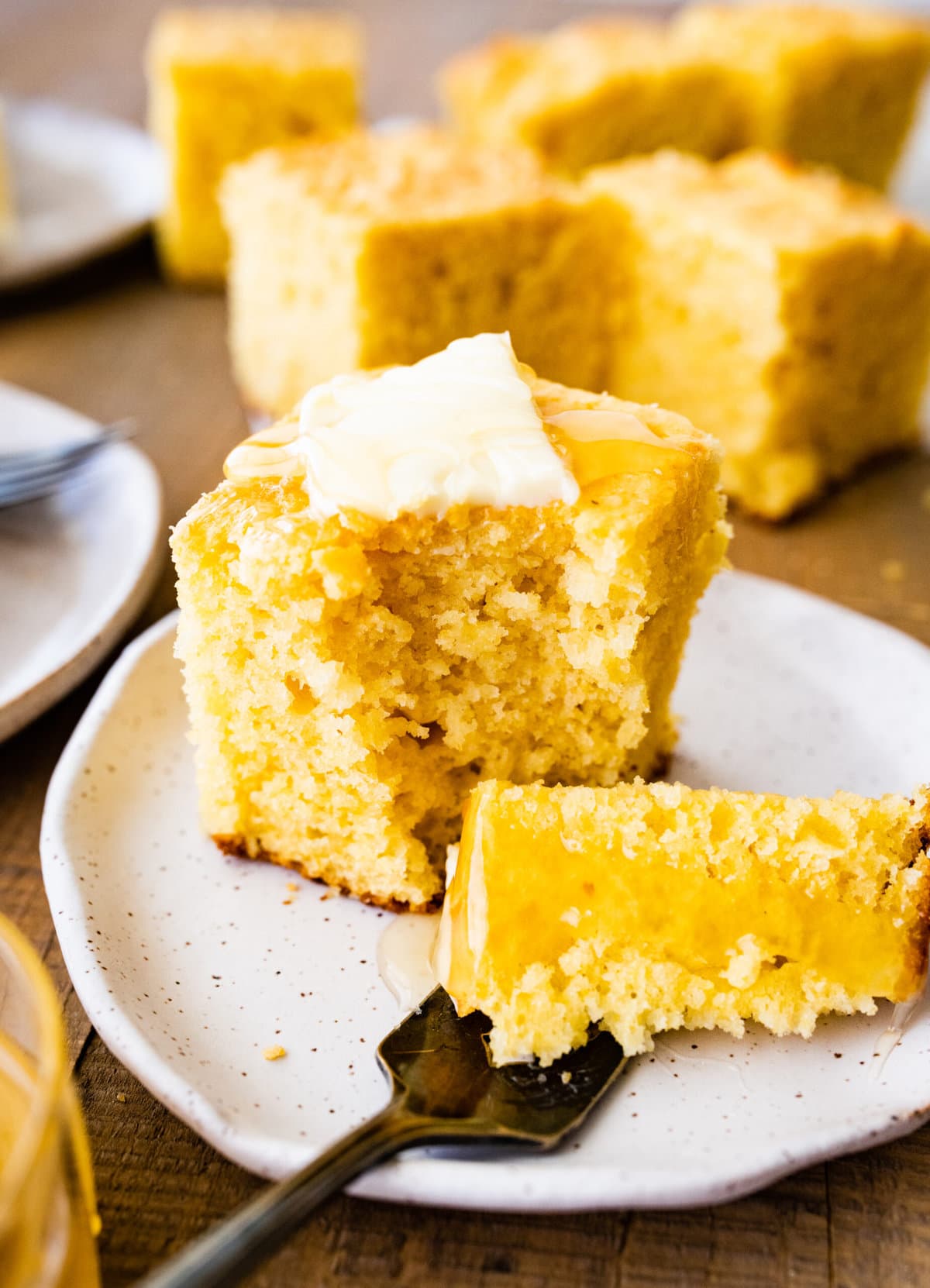 fork holding a bite of the cornbread. in the background slice of sweet cornbread and butter and honey.