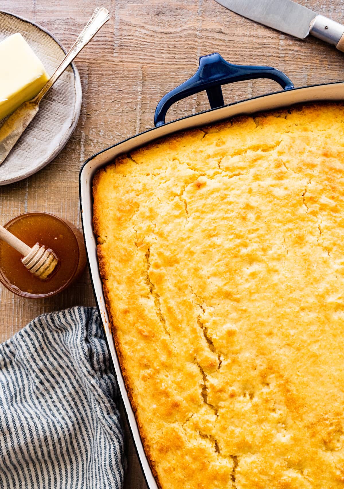 gorgeous baked cornbread in a blue and white ceramic baking dish. Small bowl of honey on the side with a blue and white dish towel.