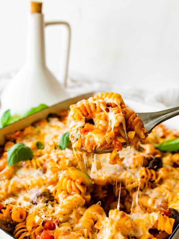 Cheesy Baked Rotini pasta after it is baked melty cheese on top with a few basil leaves. Spatula lifting up with a cheese pull.