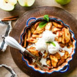 Easy baked cinnamon apples in a pie plate with blue rim. Vanilla Ice cream in the middle of apples with serving spoon. Cut apples in the background.