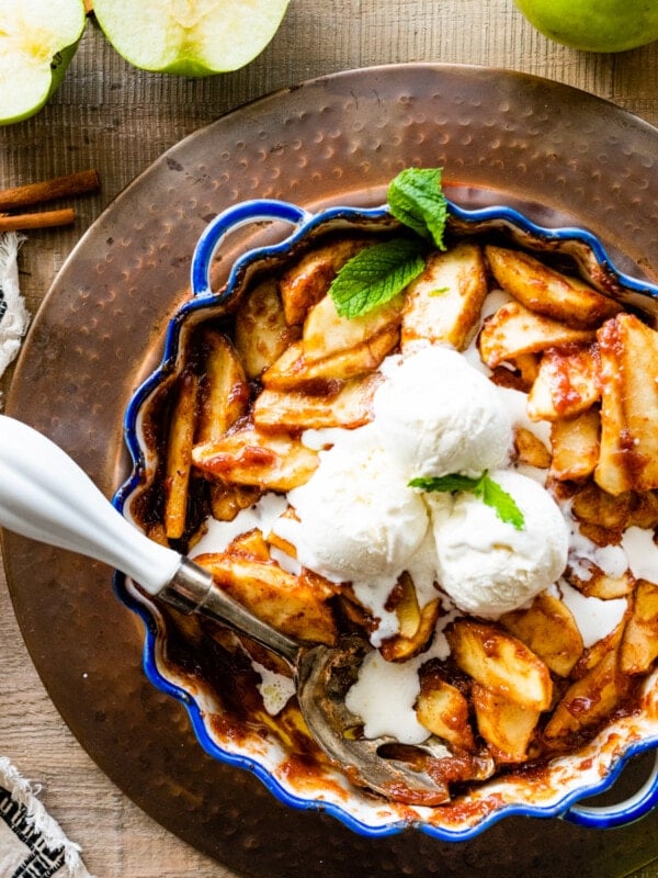 Easy baked cinnamon apples in a pie plate with blue rim. Vanilla Ice cream in the middle of apples with serving spoon. Cut apples in the background.