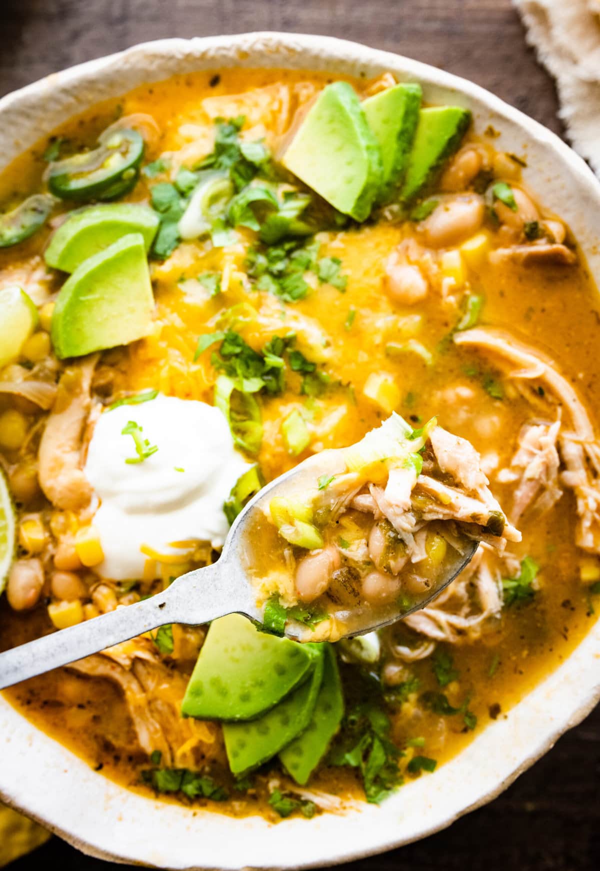 bowl of healthy white chicken chili with avocado, sour cream, lime, and cilantro as toppings. Chips in the bowl and on the side. Spoon taking a bit of the soup.