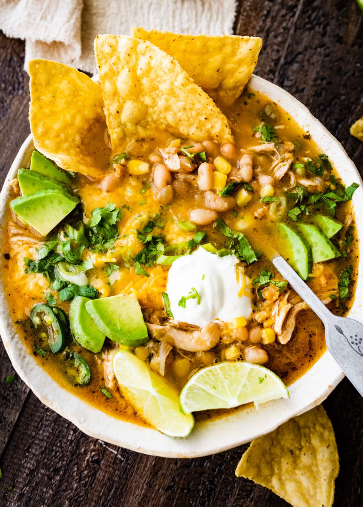 bowl of healthy white chicken chili with avocado, sour cream, lime, and cilantro as toppings. Chips in the bowl and on the side. Spoon in the bowl. 