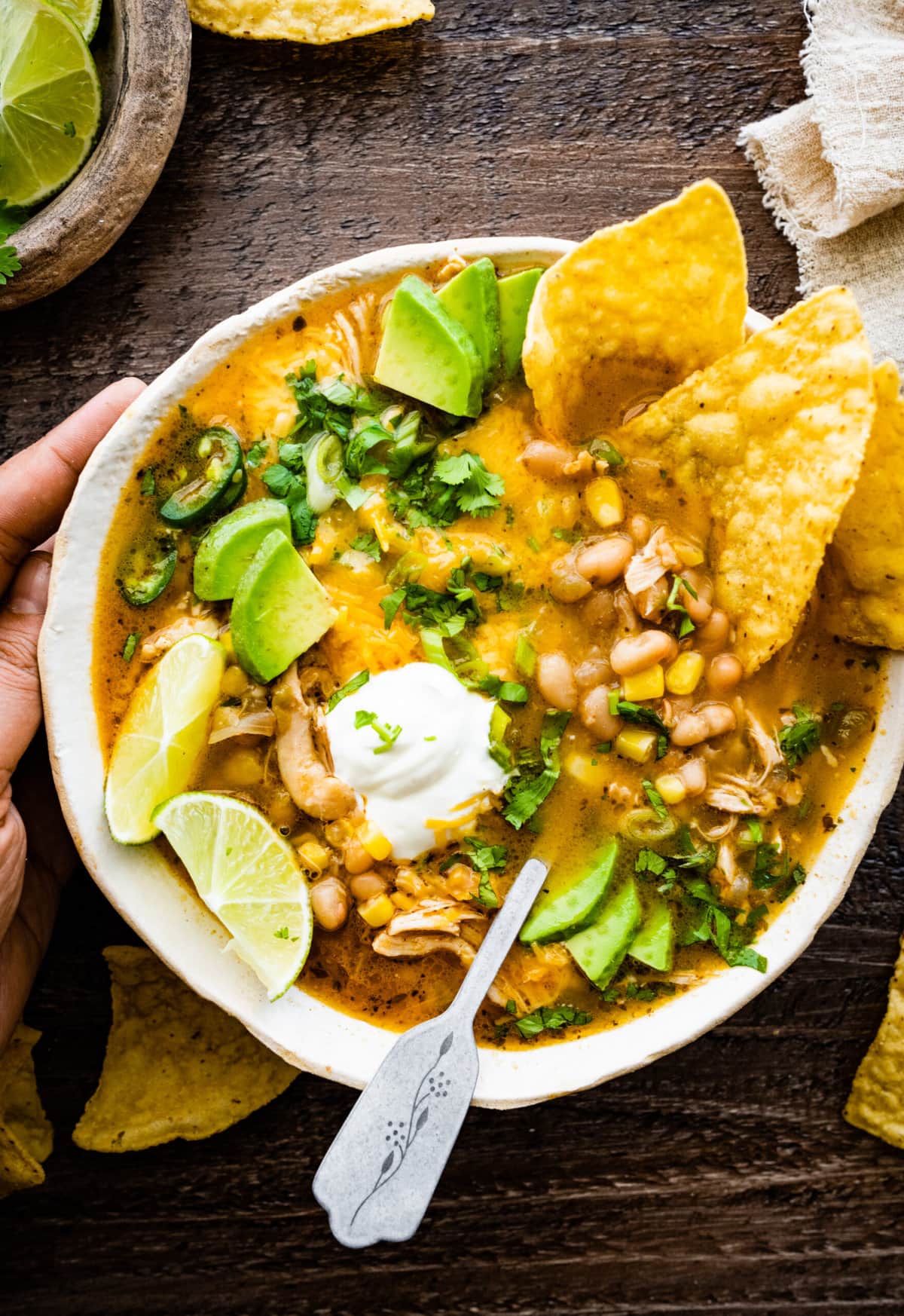 bowl of healthy white chicken chili with avocado, sour cream, lime, and cilantro as toppings. Chips in the bowl and on the side. Hands holding the side of the bowl.