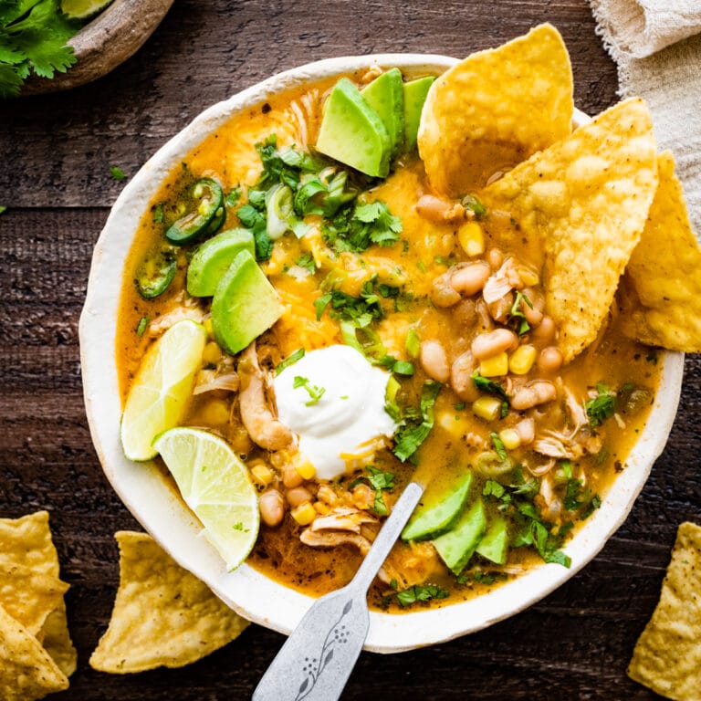 bowl of healthy white chicken chili with avocado, sour cream, lime, and cilantro as toppings. Chips in the bowl and on the side.
