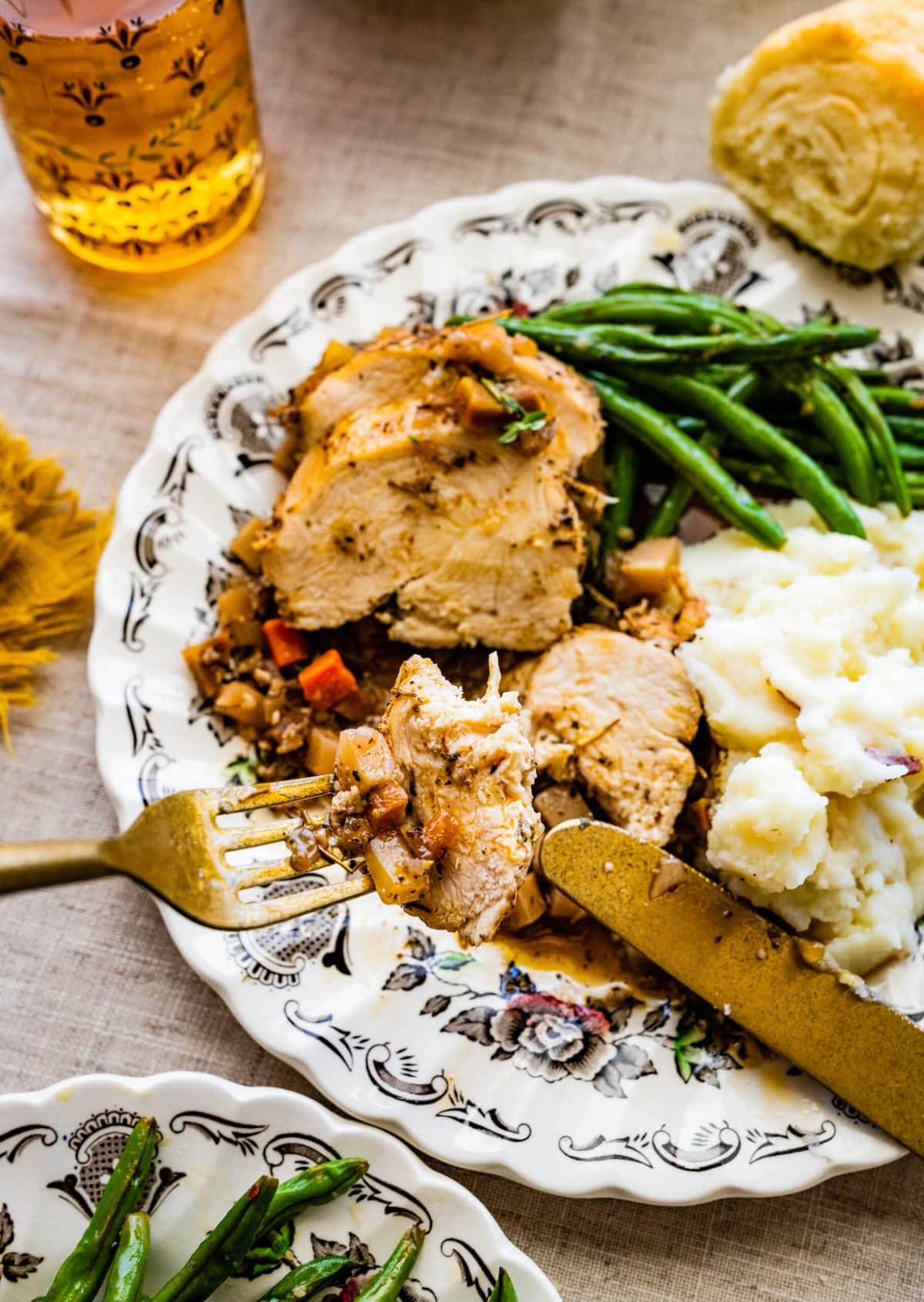 Juicy Oven Baked Chicken Breast Recipe on a plate with mashed potatoes and green beans and homemade rolls. beautiful table setting with silverware. Bite of chicken on a fork.