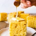 thick square of sweet cornbread on a plate with a pat of butter and honey drizzled on top.