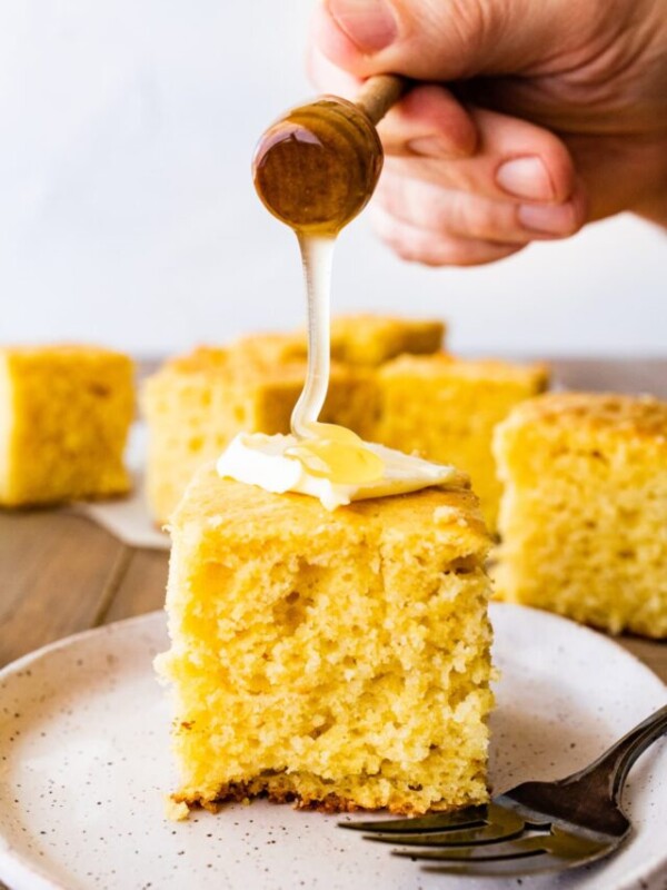 thick square of sweet cornbread on a plate with a pat of butter and honey drizzled on top.