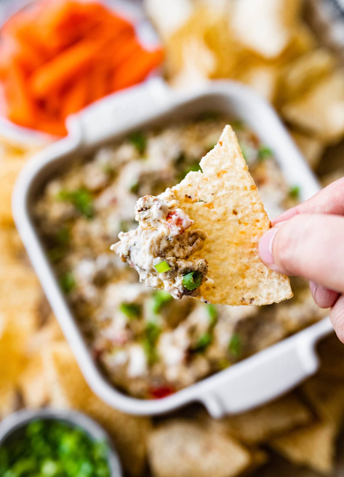 Best Jimmy Dean Sausage dip in a white serving dish with chips surrounding the dish. Hand holding chip with sausage dip on it.