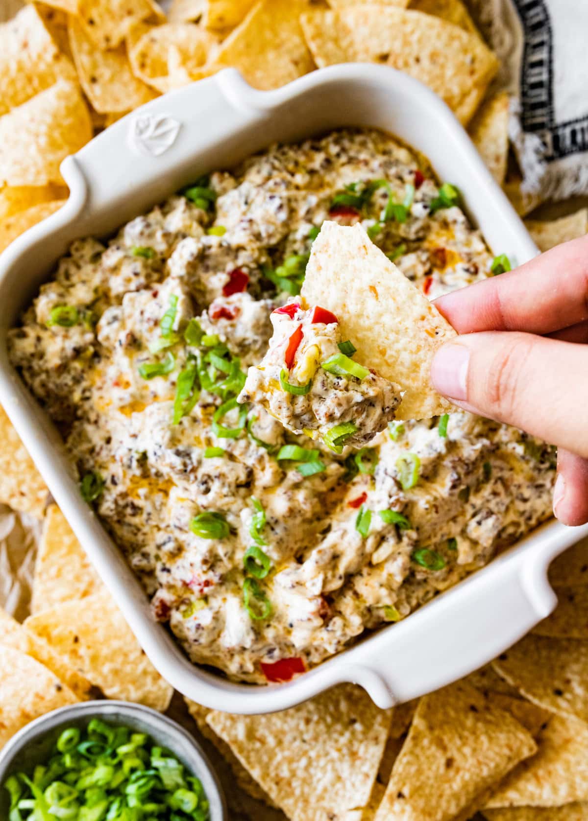 Best Jimmy Dean Sausage dip in a white serving dish with chips surrounding the dish. Hand holding chip with sausage dip on it. 