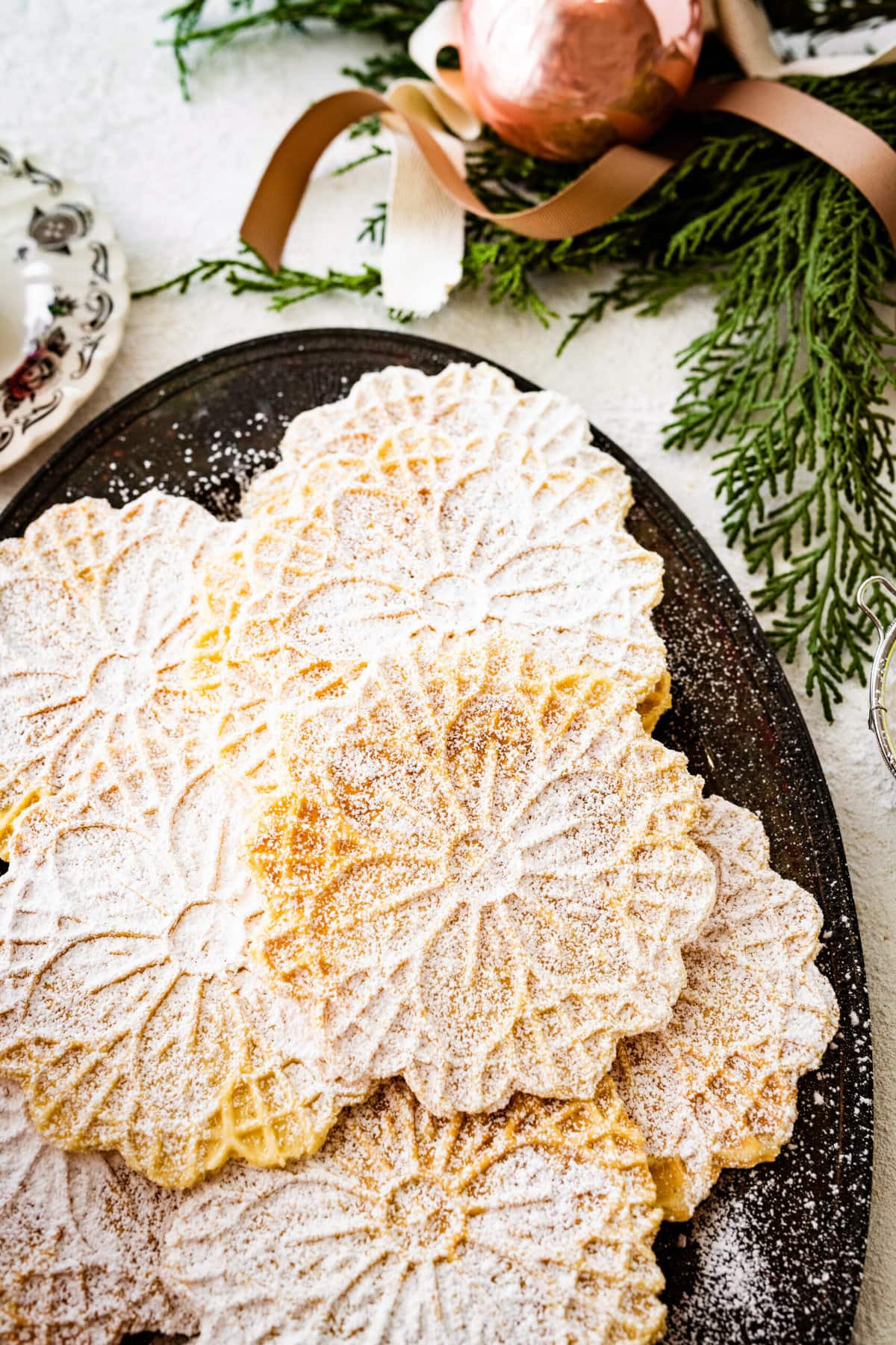 best pizzelle cookies on a black tray with powdered sugar on top. Christmas decorations all around and tea cup. 