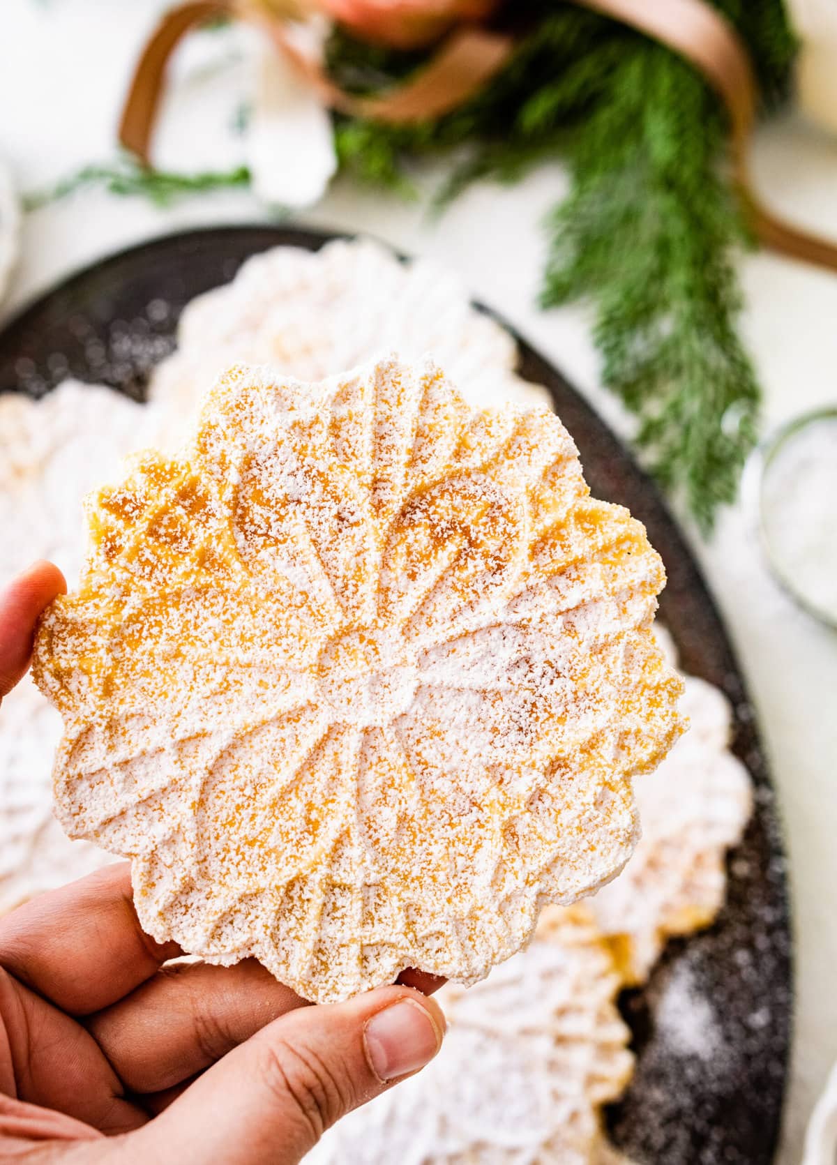 best pizzelle cookies on a black tray with powdered sugar on top. Christmas decorations all around and tea cup. Hand holding up one cookie to show pretty lace pattern.