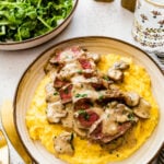 sliced steak on a plate with mushroom sauce over the top on top of a polenta. Fork and knife next to plate with green salad on the side.