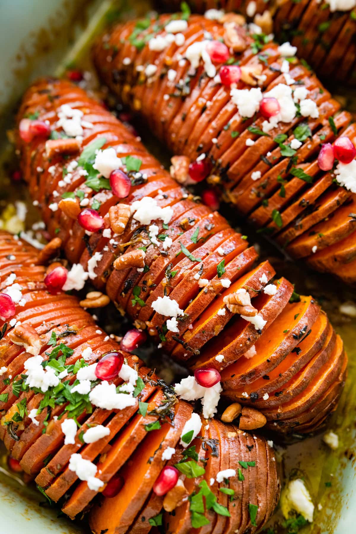 Hasselback Sweet Potatoes with Maple Syrup: potatoes after they are done baking in a teal baking dish. Add feta cheese and pomegranate seeds as toppings on the backed hassleback potatoes. Close up of the potatoes. Added hazelnuts on top.