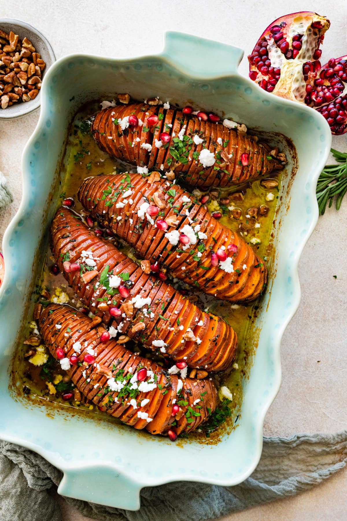 Hasselback Sweet Potatoes with Maple Syrup: potatoes after they are done baking in a teal baking dish. Add feta cheese and pomegranate seeds as toppings on the backed hassleback potatoes. Close up of the potatoes. Added hazelnuts on top.