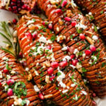 hassleback sweet potatoes on a beautiful white serving platter garnished with fresh herbs and pomegranate seeds. Close up of the the potatoes.