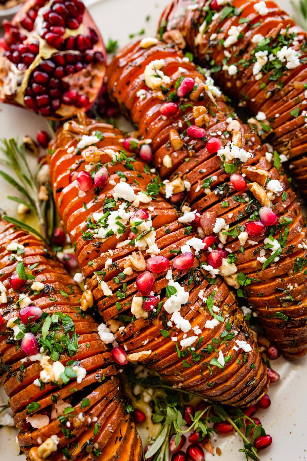 hassleback sweet potatoes on a beautiful white serving platter garnished with fresh herbs and pomegranate seeds. Close up of the the potatoes.