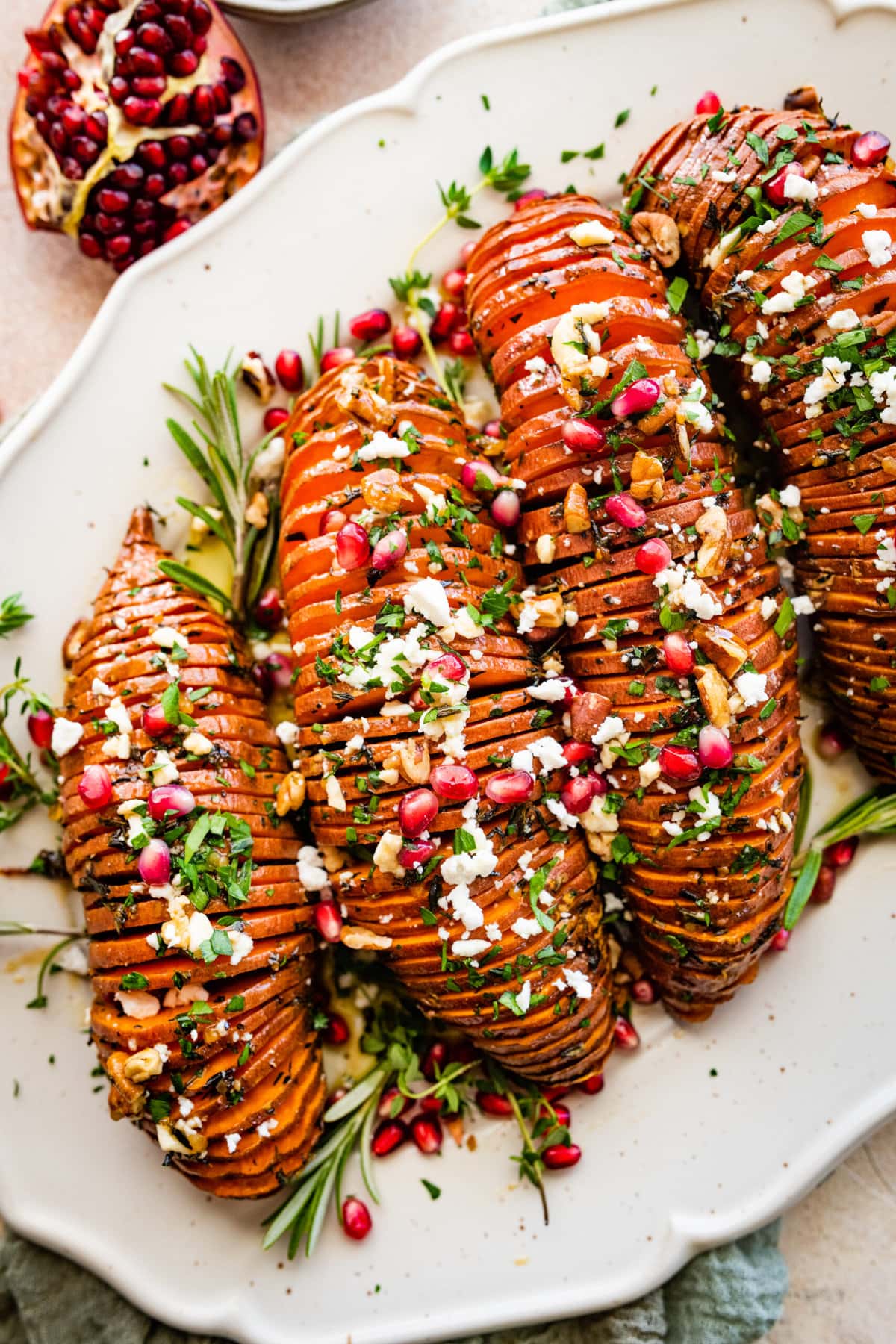 hassleback sweet potatoes on a beautiful white serving platter garnished with fresh herbs and pomegranate seeds.