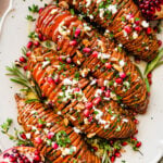 hassleback sweet potatoes on a beautiful white serving platter garnished with fresh herbs and pomegranate seeds.
