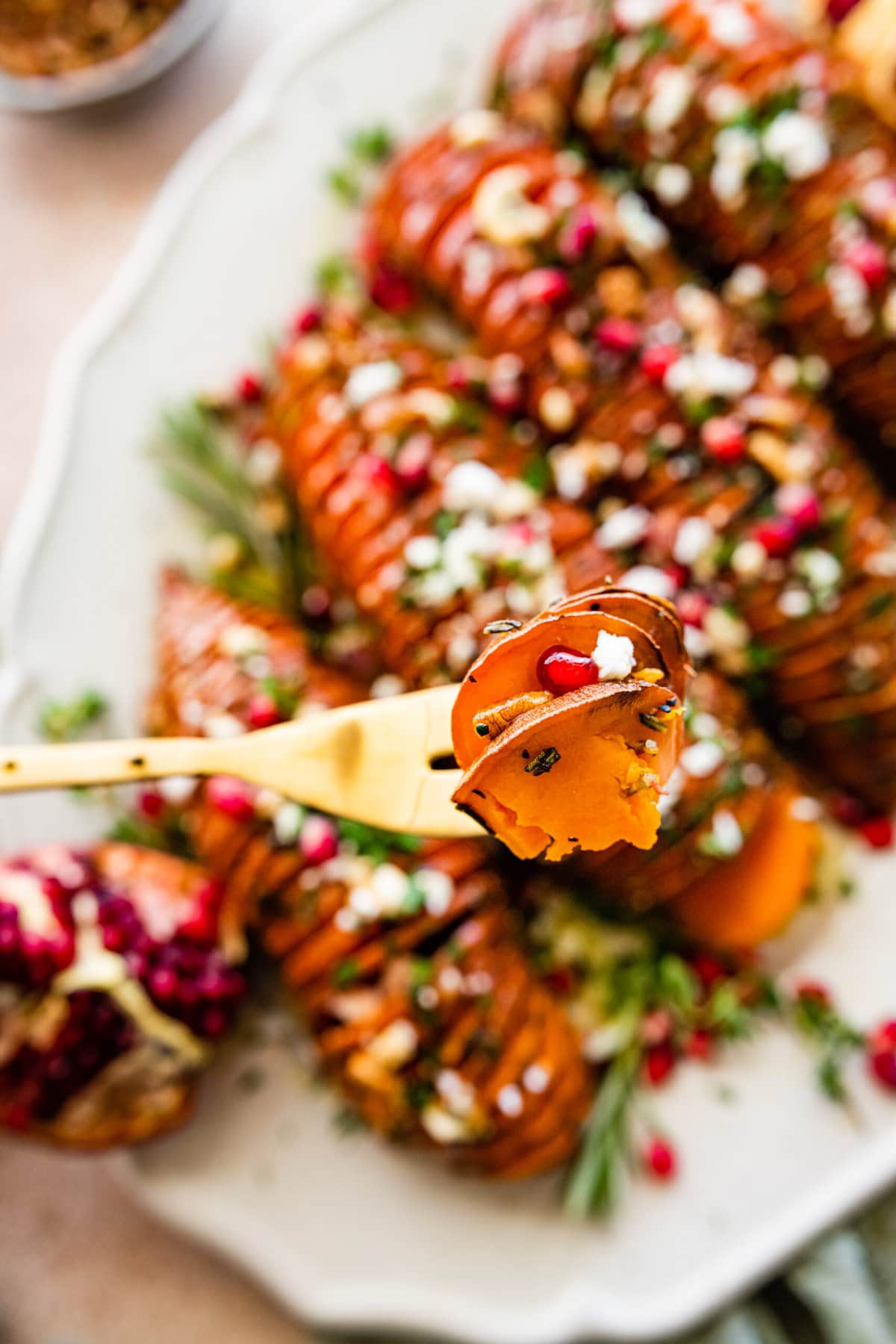 hassleback sweet potatoes on a beautiful white serving platter garnished with fresh herbs and pomegranate seeds. Bite with a fork holding it up.