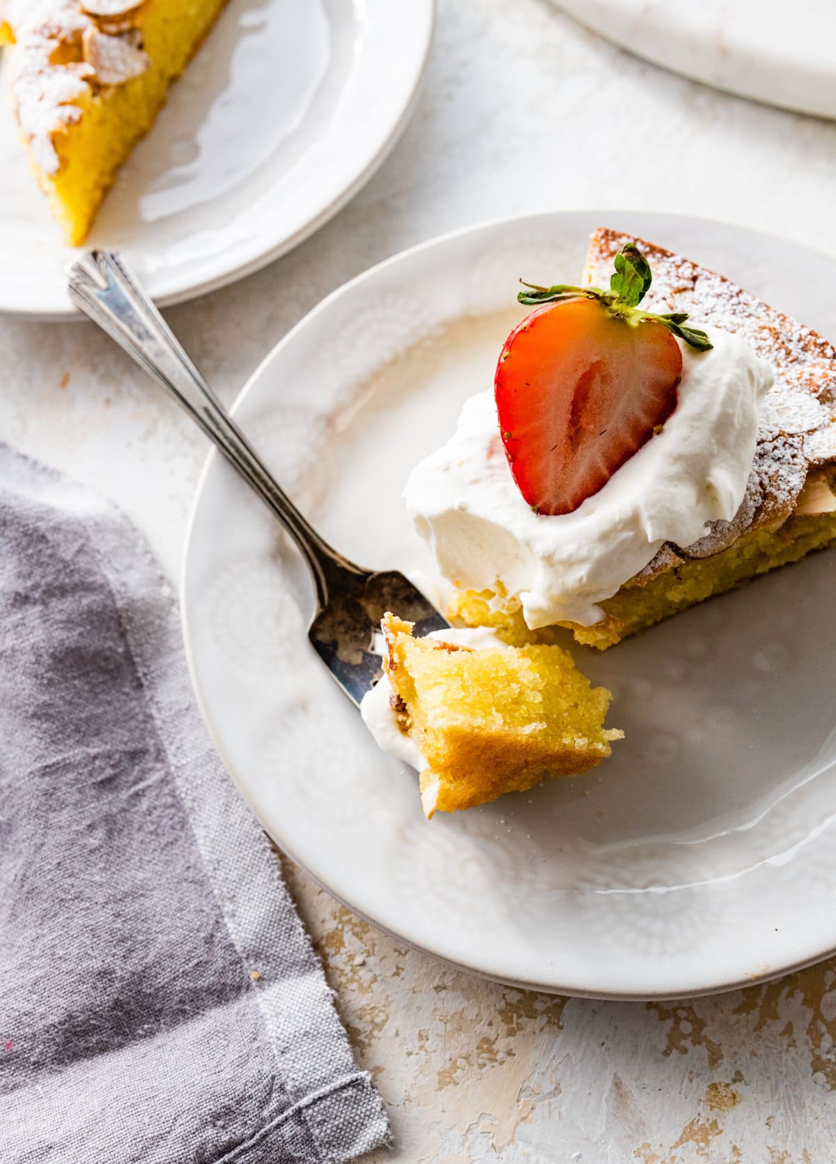 easy almond cake recipe on a plate with a fork taking a bite. Cake topped with whipped cream and strawberry.