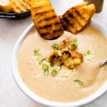 bowl of cauliflower soup with grilled bread on the side and whole pieces of roasted cauliflower on top.