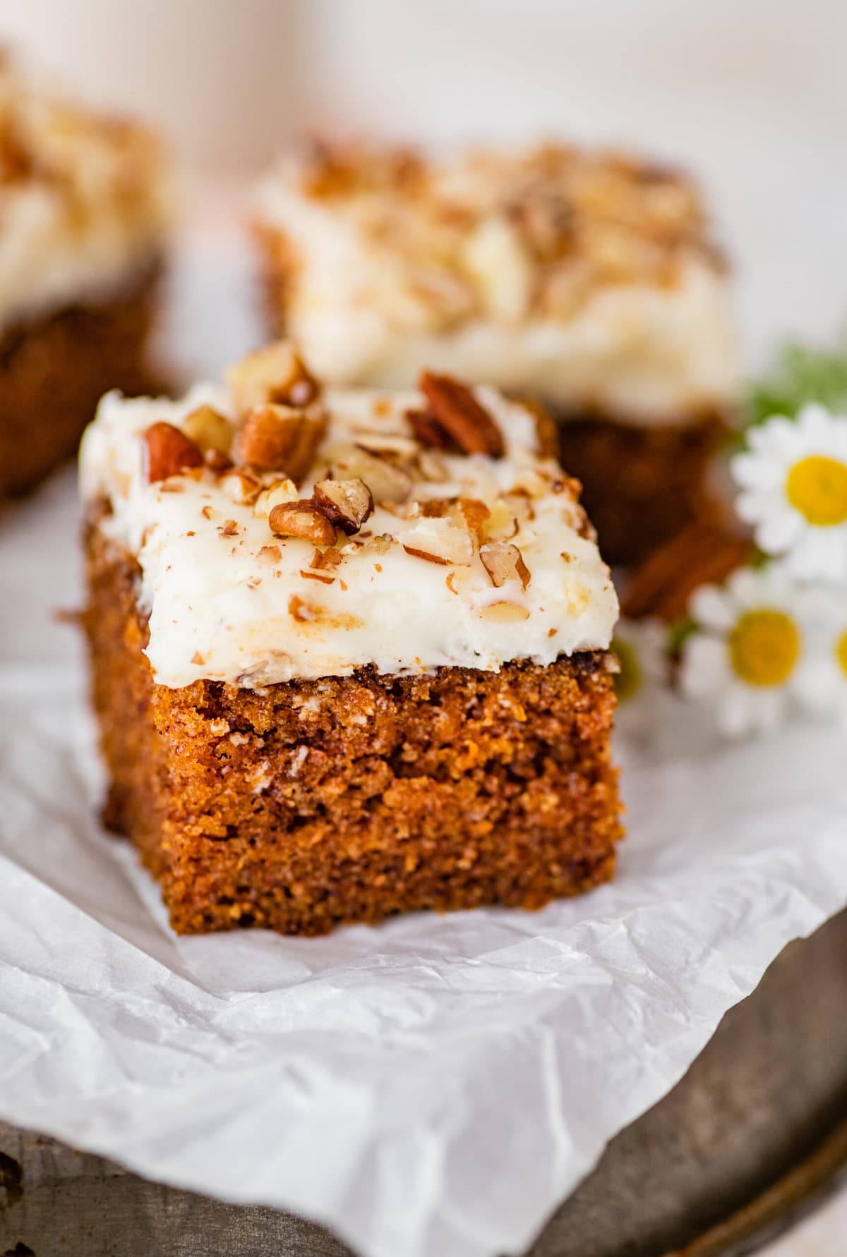 a tray of square slices of carrot cake with daisy flowers in the background.