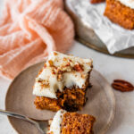 a square slice of carrot cake with cream cheese frosting on a plate. bite taken out of the cake with a fork.