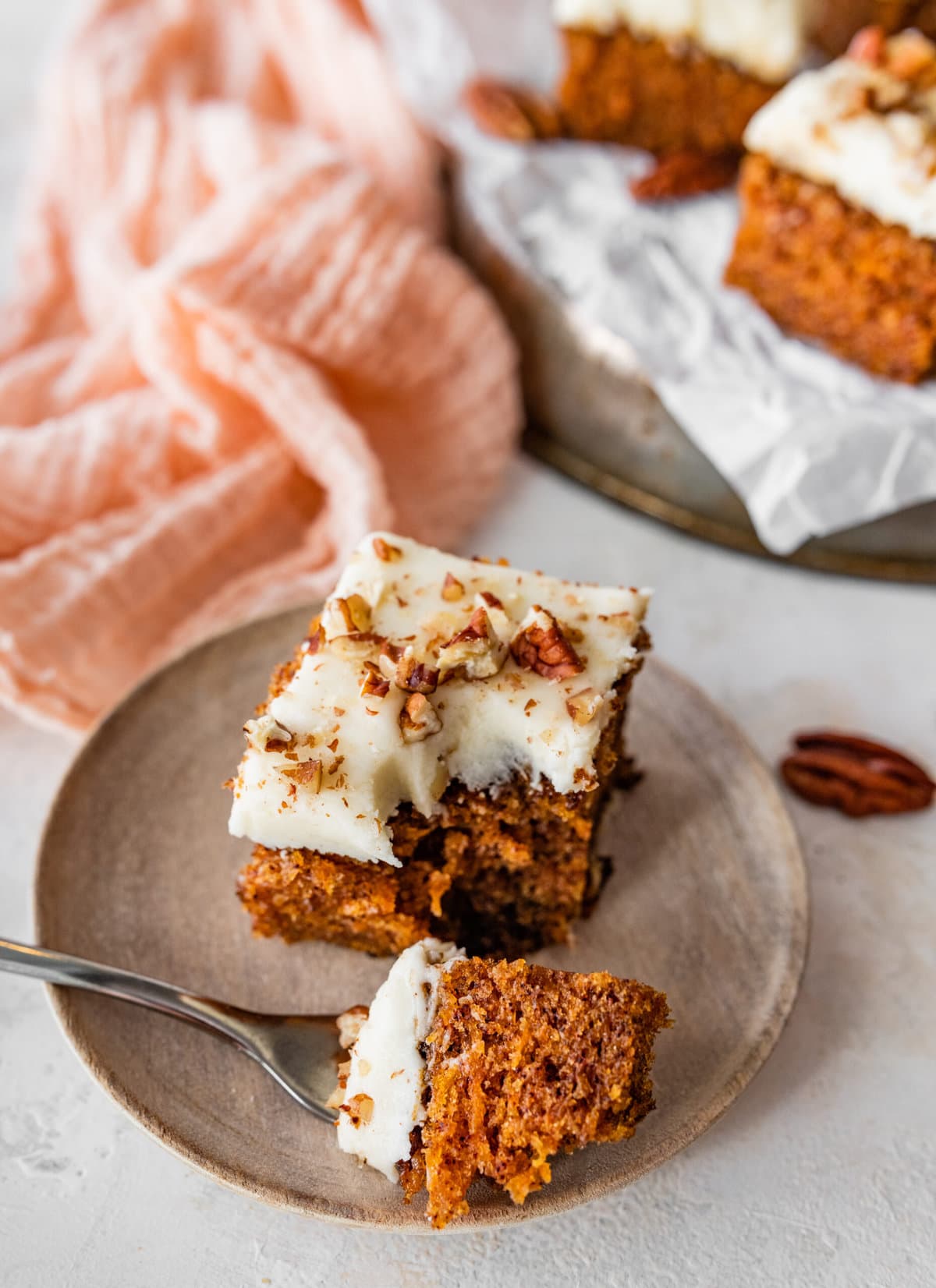 a square slice of carrot cake with cream cheese frosting on a plate. bite taken out of the cake with a fork.