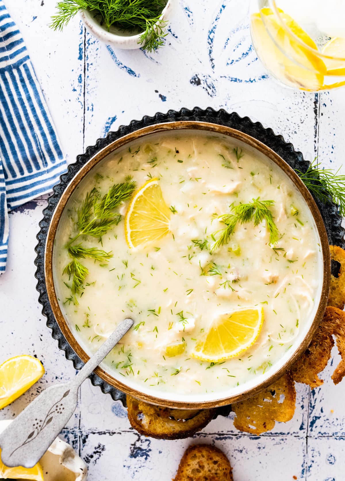 Avgolemono soup on a blue and white backdrop with fresh dill as garnish. Fresh lemon as a side.
