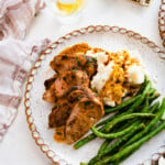 pork tenderloin slices on a plate with green beans and mashed potatoes. pan juices over the top.