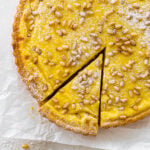 finished torta della nonna with pine nuts on top after baking. a light dusting of powdered sugar. One slice taken out of the torta. cover photo.
