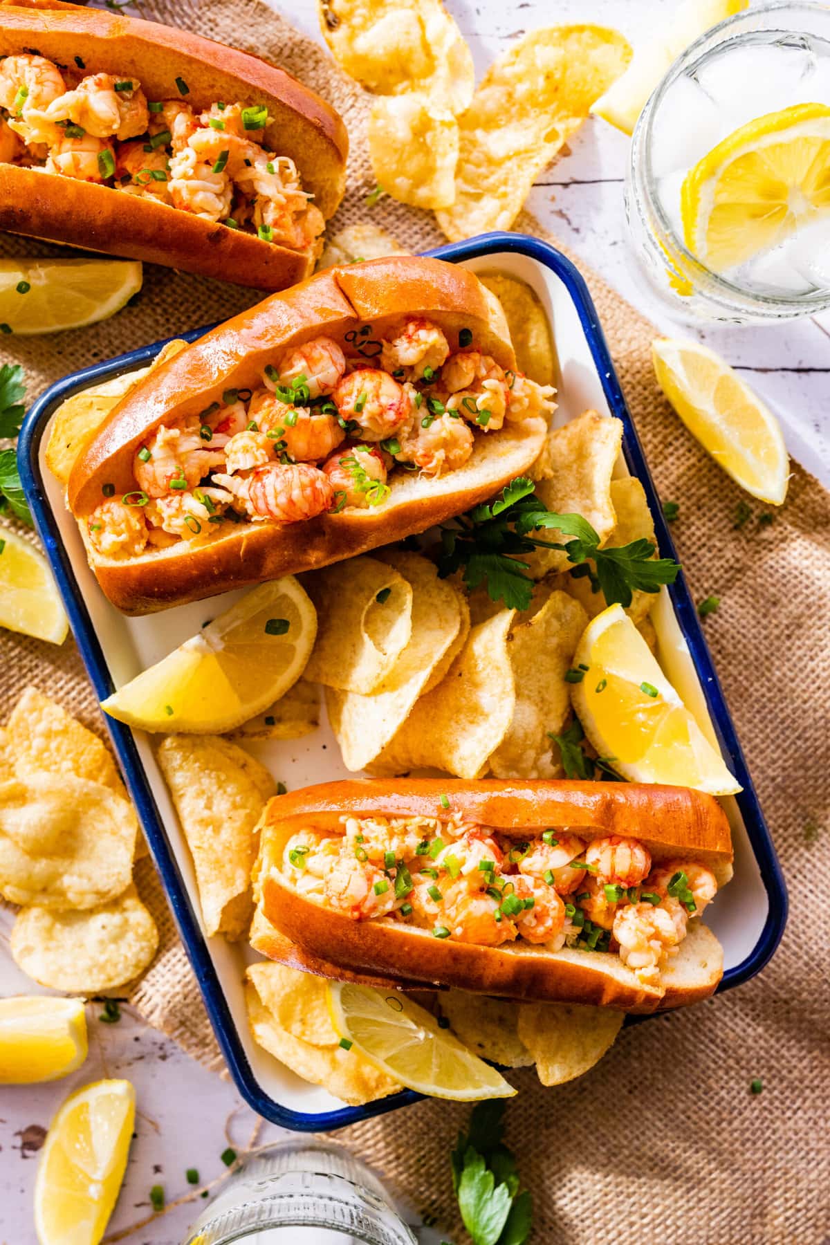 Two langostino lobster rolls on a plate with chips and lemon wedges.