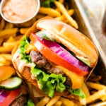 best hamburger recipe ever perfect juicy burger- hand holding the burger ready to eat. Fries and sauce in the background.
