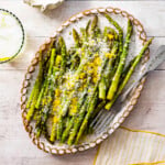 asparagus finished cooking on a white platter with freshly grated parmigiano and lemon zest on it. Fork on plate. water glass with lemon on the side.