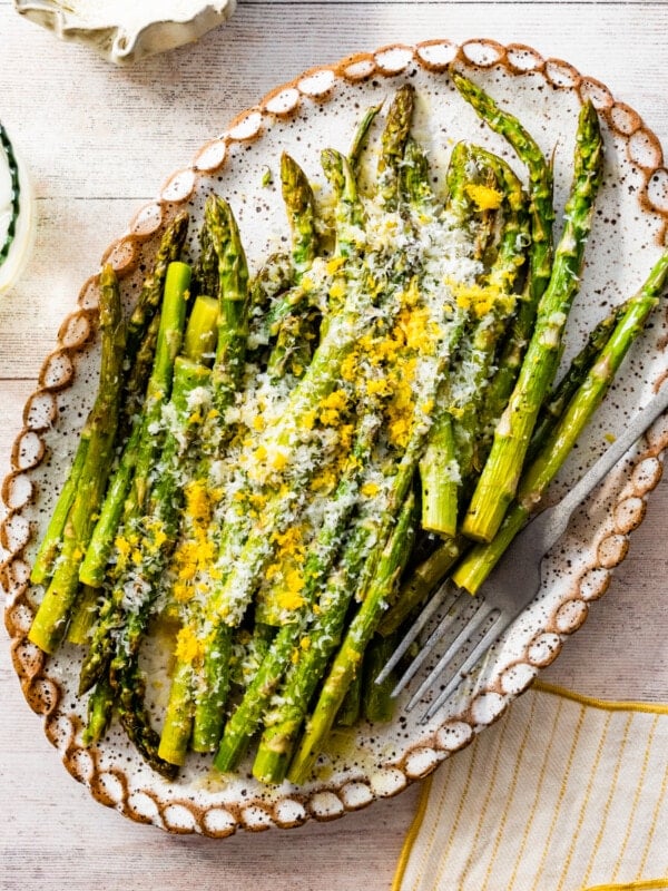 asparagus finished cooking on a white platter with freshly grated parmigiano and lemon zest on it. Fork on plate. water glass with lemon on the side.