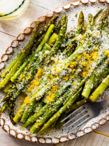 asparagus finished cooking on a white platter with freshly grated parmigiano and lemon zest on it. Fork on plate. water glass with lemon on the side.