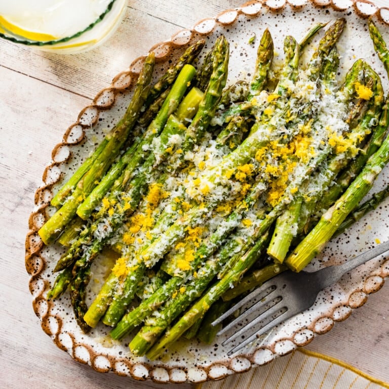 asparagus finished cooking on a white platter with freshly grated parmigiano and lemon zest on it. Fork on plate. water glass with lemon on the side.