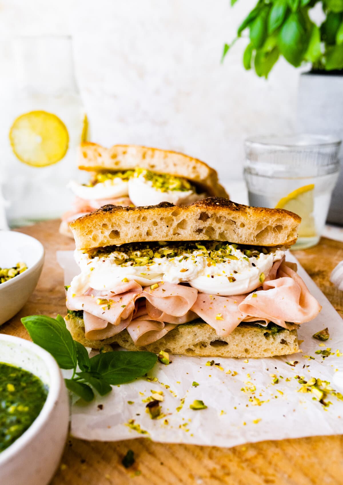 Assembled Mortadella Sandwich Recipe with Pistachio Pesto and Burrata with pesto on the side. Ready to eat. Lemon water glasses in the background.