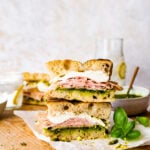 Assembled Mortadella Sandwich Recipe with Pistachio Pesto and Burrata with pesto on the side. Sandwiches stacked on one another to see the layers on the inside. Ready to eat. Lemon water glasses in the background.