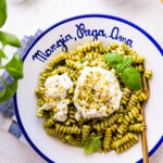 platted bowl of pasta with burrata cheese crushed pistachios on the top. The bowl is white and in blue is written the Italian phrase- "Mangia, Prega, Ama". N;ue and white napkin and fork in plate.
