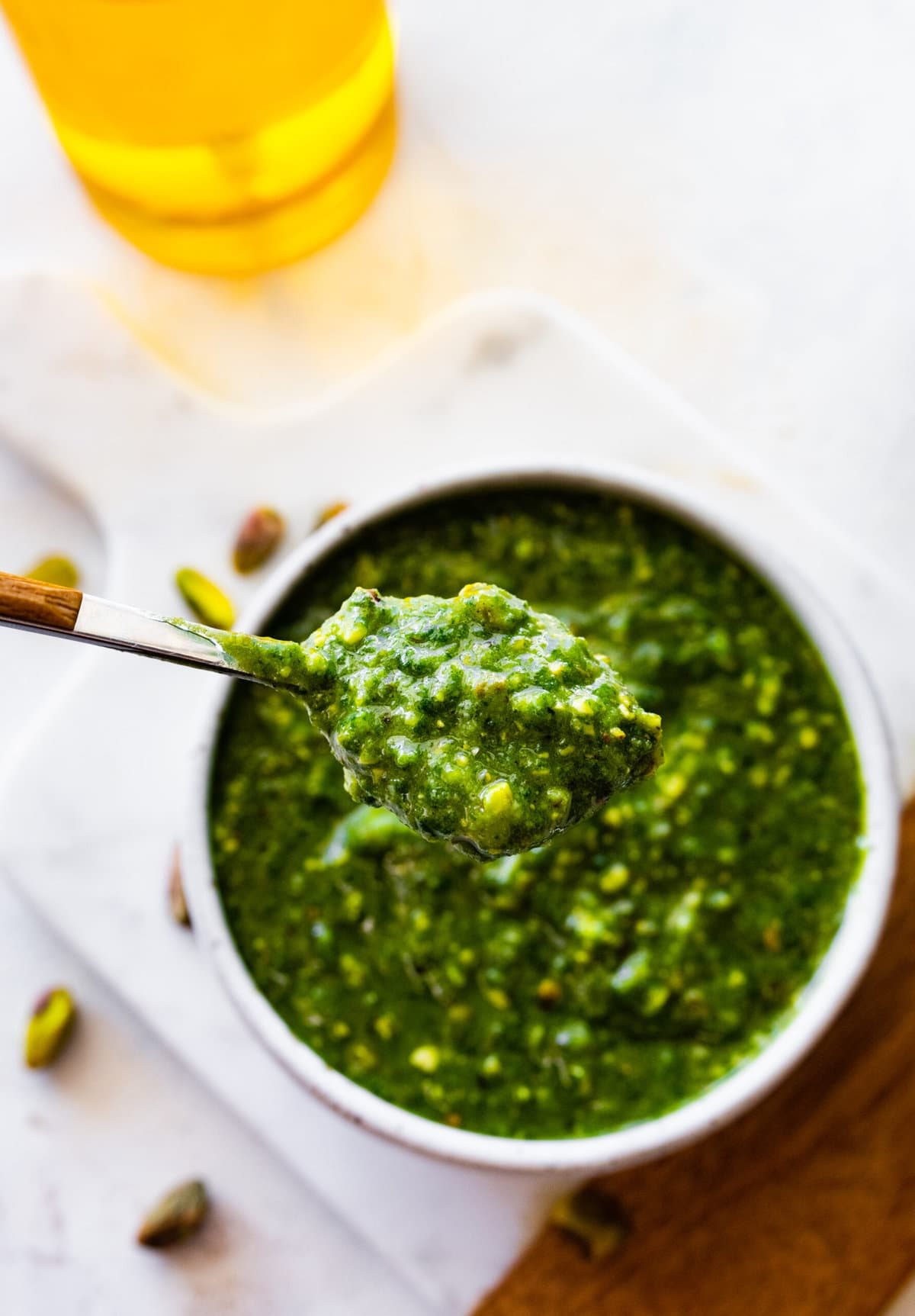 pistachio pesto in a bowl with a spoon in it on a marble and wood cutting board. Spoon holding up a spoonful of the pistachio pesto.