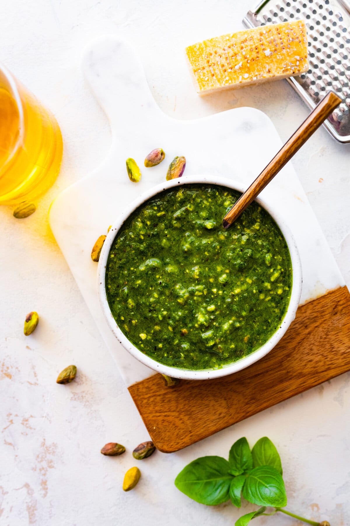 pistachio pesto in a bowl with a spoon in it on a marble and wood cutting board.