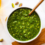 pistachio pesto in a bowl with a spoon in it on a marble and wood cutting board.