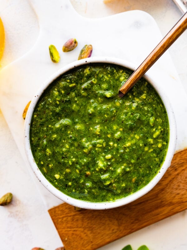 pistachio pesto in a bowl with a spoon in it on a marble and wood cutting board.